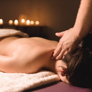 Men's hands make a therapeutic neck massage for a girl lying on a massage couch in a massage spa with dark lighting. Close-up. Dark Key.